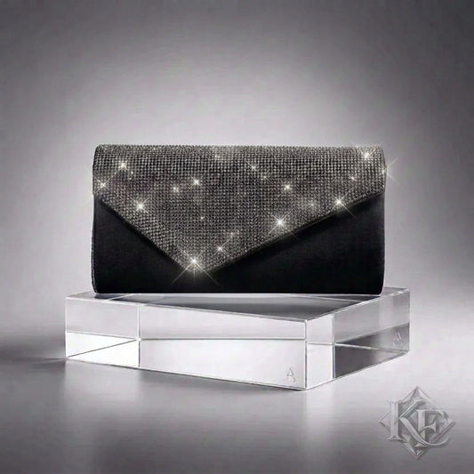 A black clutch bag with a silver rhinestone-embellished triangular flap, displayed on a clear acrylic block against a gray gradient background.