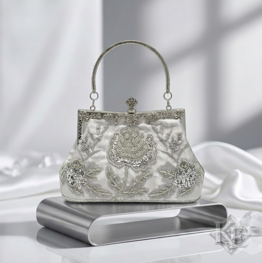 An ornate silver evening clutch bag with intricate floral embroidery and crystal embellishments, displayed on a metallic stand against a draped white fabric background.