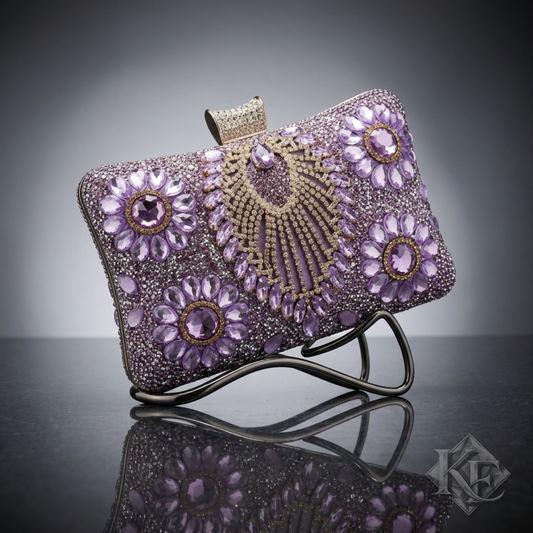 A rectangular evening clutch purse covered in lavender and silver rhinestones, featuring a central gold-tone beaded fan design and floral motifs, with a small gold clasp at the top, shown on a light gray reflective surface.